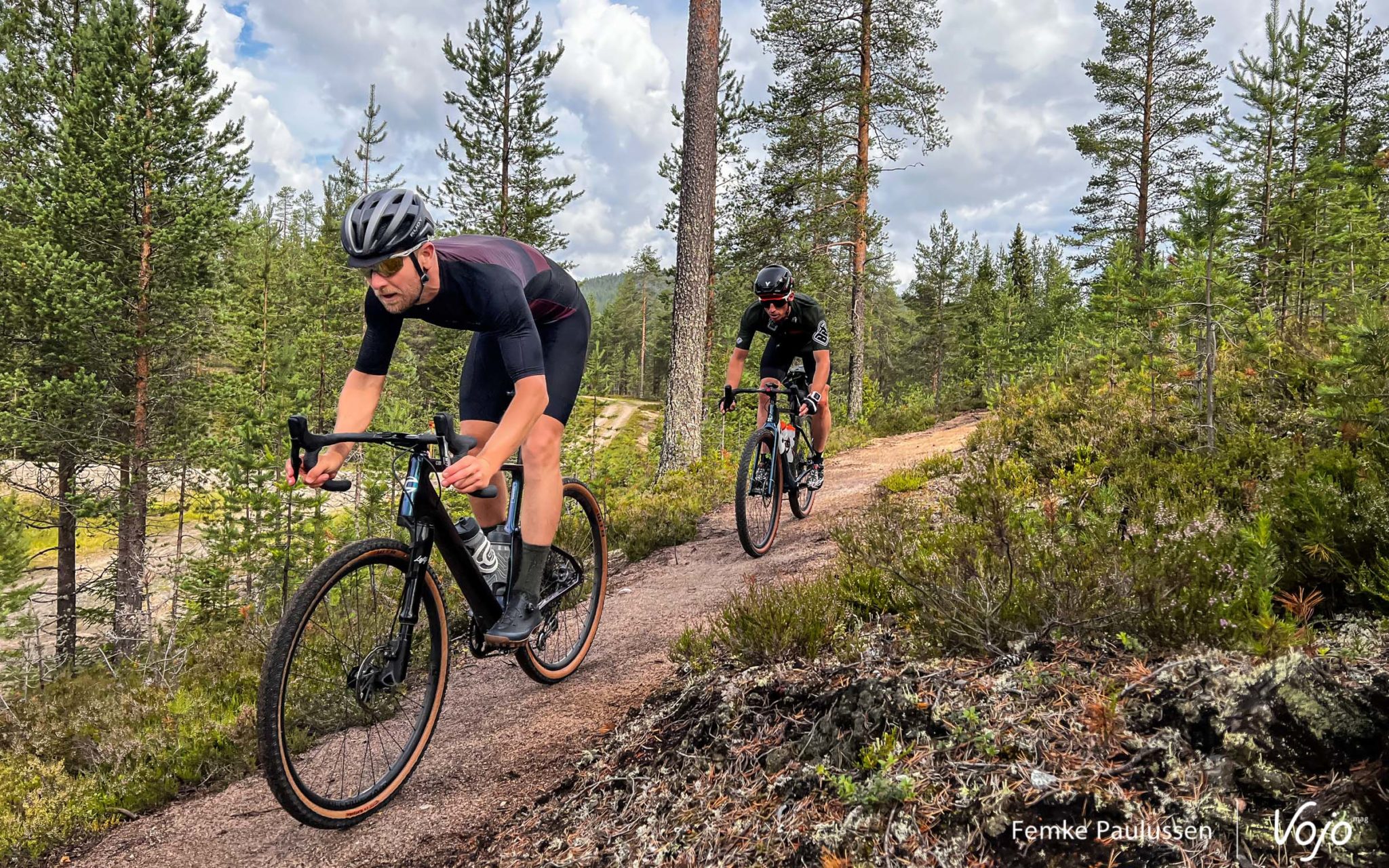 Deux hommes en gravel en descente, sur un petit sentier lisse dans un paysage scandinave (mousses, lichens et grands conifères espacés)